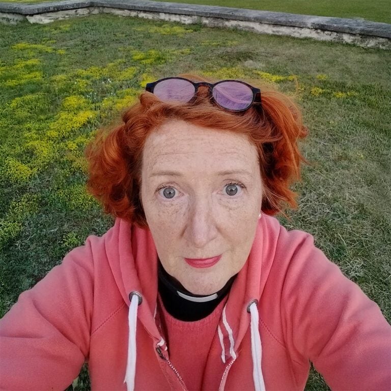 Pappi Jaana Rantala vaaleanpunaisessa hupparissa niityllä.